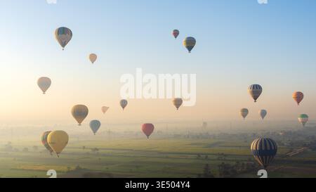 Dichte Gruppe von bunten Heißluftballons, die bei Sonnenaufgang über grüne Felder und Palmen schweben, in Luxor, Ägypten Stockfoto