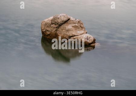 Ein großer, strukturierter Felsen liegt in sehr stilem Wasser. Die klare Reflexion des Gesteins ist auf der glatten Wasseroberfläche sichtbar. Die Beleuchtung ist weich und gleichmäßig Stockfoto