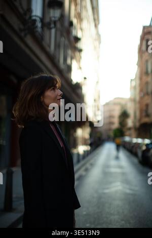 Eine junge Frau mit braunen Haaren, die einen schwarzen Blazer trägt, steht auf einer Straße der Stadt und blickt auf Gebäude, die von Sonnenlicht im Hintergrund beleuchtet werden Stockfoto