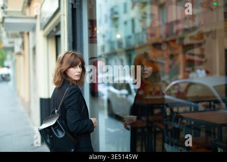 Eine weibliche Figur mit braunem Haar in schwarzer Kleidung steht vor einem Café und blickt auf ihr Spiegelbild im Glasfenster, in dem die urbane Straßenszene sichtbar ist Stockfoto