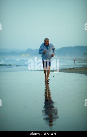 Senior Mann joggt bei Sonnenaufgang am Strand entlang, trägt einen hellblauen Hoodie und Shorts, mit Reflexen im nassen Sand und den Wellen Stockfoto