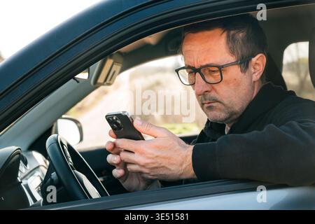 Reifer Mann mit Brille sitzt im Auto und überprüft das Smartphone, während er nach einer Fahrzeugpanne auf den Pannendienst wartet, alltäglichen Pendelverkehr und mobil u Stockfoto