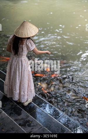 Asiatische Frau in konischem Hut und schickem Kleid genießt Süßwasserkarpfen im Teich im Park. Stockfoto