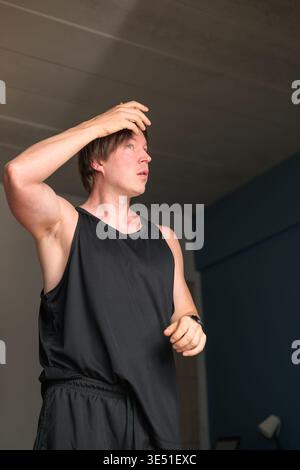 Ein Mann im mittleren Erwachsenenalter steht in einem minimalistischen Schlafzimmer mit einem schockierten Ausdruck, der seine Stirn berührt. Die Inneneinrichtung verfügt über einen blauen Akzent mit Stockfoto