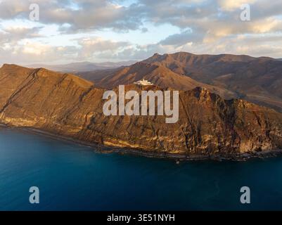 Blick aus der Vogelperspektive auf zerklüftete, sonnenverwöhnte Berge, die auf das türkisfarbene Meer treffen, gekrönt von einem unberührten weißen Leuchtturm, Fuerteventura, Kanarischen Inseln, Spanien. Stockfoto