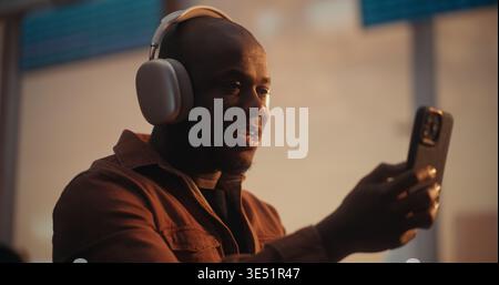 Black man trägt stylische Silver Over Ear Kopfhörer und Overshirt und nimmt Videochat auf dem Smartphone auf. In der Airport Departure Lounge sitzen, man lächelt und Reisebegeisterung mit Lieben teilen. Stockfoto
