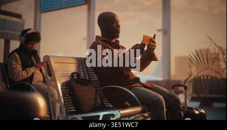 Schwarzer Mann, der Laptoptasche und Gepäck trägt, betritt den geräumigen Wartebereich am Flughafen mit Fenstern mit Blick auf den Airfield. Der Mann verwendet das Smartphone, um den Pass zu scannen und die Flugdaten und Gate-Informationen zu überprüfen. Stockfoto