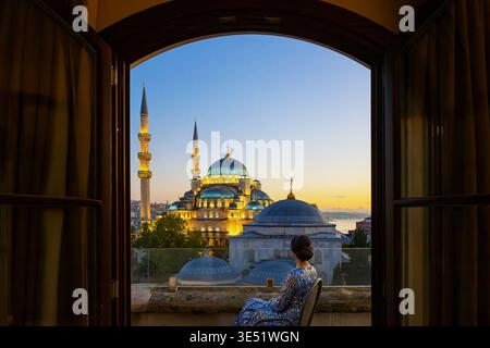 Eine attraktive Frau in nahöstlicher Kleidung sitzt auf einem Balkon bei Sonnenuntergang mit Blick auf die New Yeni Moschee und die Stadt Istanbul, Türkei. Stockfoto