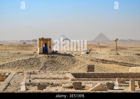 Die Ruinen der ägyptischen Nekropole von Sakkara auf dem Gizeh-Plateau mit Arbeitern, die eine Pause im Schatten machen, und der Komplex der Großen Pyramiden im Blick dahinter. Stockfoto