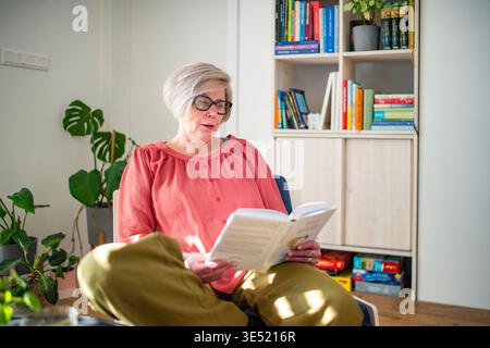 Lächelnde ältere Frau, die zu Hause Buch liest. Stockfoto