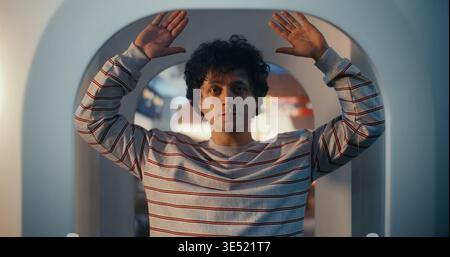 Junger Mann mit lockigem Haar in gestreiftem Sweatshirt steht im modernen Millimeter Wave Body Scanner. Guy hält die Hände hoch, wie es von den Sicherheitsprotokollen angewiesen wurde. Standardverfahren für die Luftsicherheit. Stockfoto