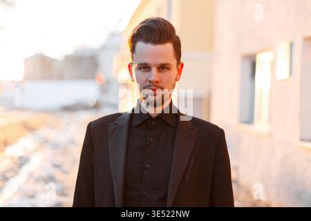 Stilvoller eleganter Geschäftsmann in modischer Kleidung mit schwarzem Blazer und Hemd, der an einem Wintertag in der Stadt bei Sonnenuntergang spaziert Stockfoto