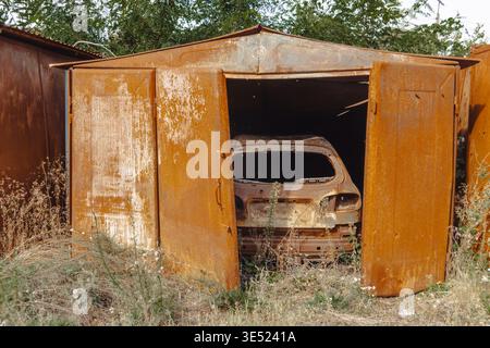 Verlassene und verbrannte rostige Metallgarage mit einer zerstörten Autoschale. Die Folgen von Militäraktionen und Feuer in einem Konfliktgebiet Stockfoto