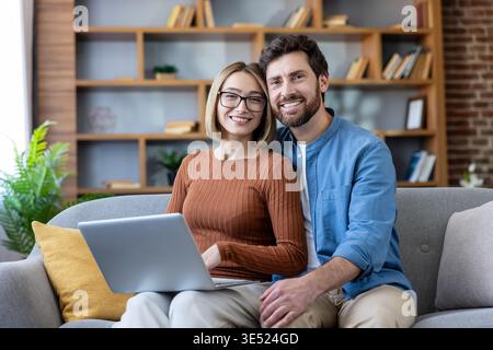 Lächelndes Paar, das auf einem Sofa in ihrem Wohnzimmer sitzt und den Betrachter ansieht, während er mit einem Laptop interagiert, repräsentiert Verbindung und modernen Lebensstil Stockfoto