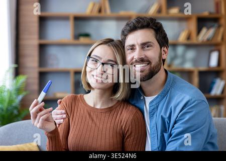 Junge lächelnde Paare, die sich auf einem Sofa umarmen, Glück und Aufregung teilen, nachdem sie ein positives Schwangerschaftstestergebnis erhalten haben, und freuen sich darauf, ihre Familie zu gründen Stockfoto