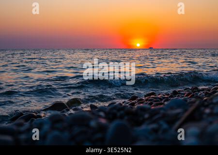 Ozeanwellen brechen an einem Kiesstrand unter einem leuchtend orangen Sonnenuntergang. Sonnenuntergang am Meer, Küstenwellen, Naturhintergrund Stockfoto