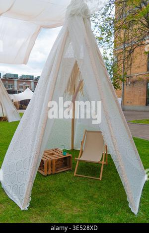 Gemütliches weißes Tipi-Zelt mit Spitze auf grünem Rasen mit hölzernem Loungesessel und rustikalem Kistentisch, perfekt für eine Lounge im Freien oder eine Veranstaltung Stockfoto