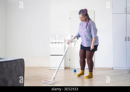 Fröhliche Frau mit Dreadlocks mit einem modernen Staubsauger in einem hellen Wohnzimmer. Stockfoto