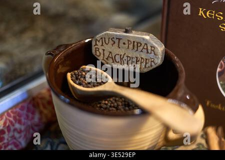 Nahaufnahme von ganzen schwarzen Pfefferkörnern in einem rustikalen zweifarbigen Keramikbehälter mit einem Holzlöffel darauf. Ein handgeschnitztes Holz Stockfoto