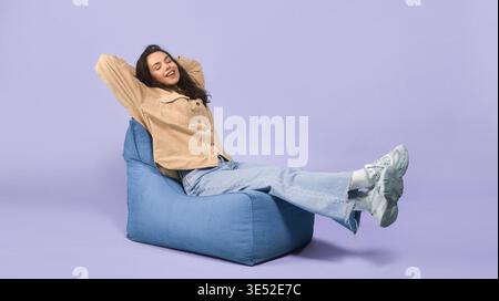 Frau sitzt auf einem blauen Sitzsessel und entspannt sich in einem hellvioletten Raum Stockfoto