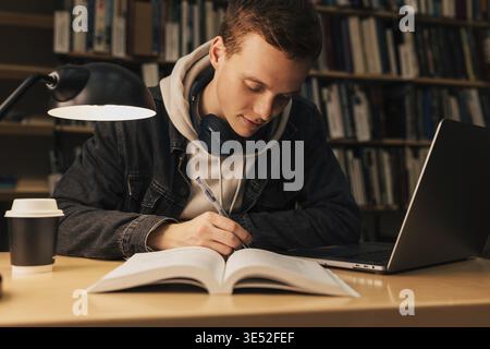 Junger Mann, der am Schreibtisch mit Laptop in der Bibliothek schrieb Stockfoto