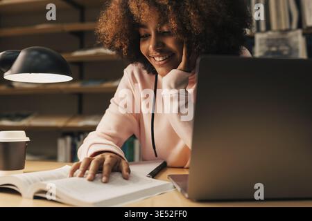 Lächelnder Student, der spät in der Nacht studiert. Junge Frau, die Buch und Laptop an der Bücherei benutzt Stockfoto