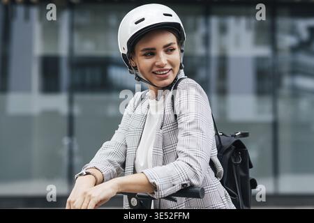 Junge Geschäftsfrau mit Elektroroller, die wegschaut. Fröhliche Frau im Schutzhelm mit Rucksack im Freien Stockfoto