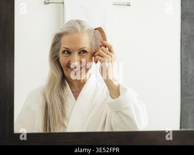 Eine Frau, die einen Holzkamm im Badezimmer hält. Eine alte Frau kämmt ihr Haar, während sie in einen Spiegel blickt Stockfoto
