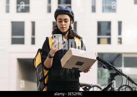 Lieferung Frau im Fahrradhelm mit einem Karton, der Informationen auf dem Handy überprüft Stockfoto