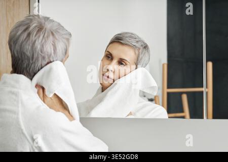 Reife Frau mit kurzen grauen Haaren, die ihre Spiegelung ansieht Im Badezimmerspiegel Stockfoto