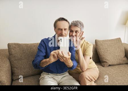 Seniorenpaar, das Videoanrufe macht, während sie auf einem Sofa im Wohnzimmer sitzt Stockfoto