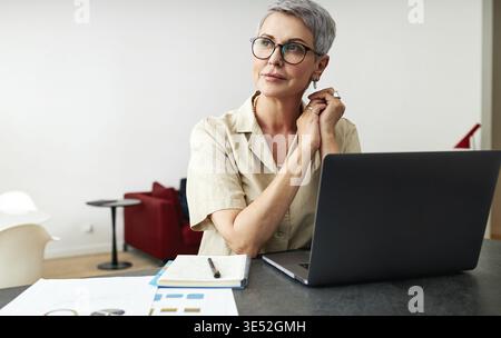 Nachdenkliche reife Frau mit Brille, die zu Hause am Schreibtisch sitzt und wegschaut Stockfoto