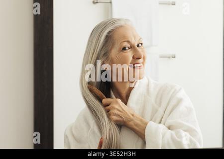 Lächelnde Frau, die ihr langes graues Haar vor einem Spiegel kämmt Stockfoto