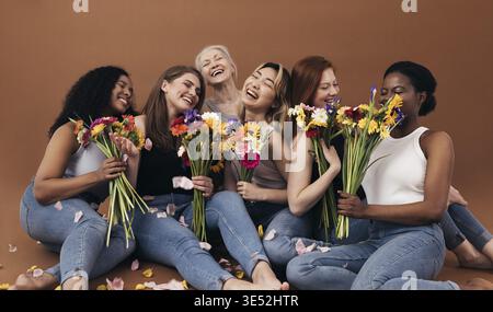 Sechs Frauen unterschiedlichen Alters und Körpertypen halten Blumensträuße, während sie im Studio vor einem braunen Hintergrund sitzen Stockfoto