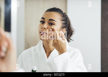 Schöne Frau im Bademantel Anwendung Anti-Aging-Creme auf ihre Wange Stockfoto