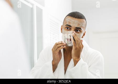 Junger Mann mit weißer Bademantel, die Gesichtsmaske im Badezimmer Stockfoto