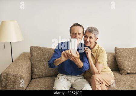 Seniorenpaar in Kasualen zu Hause auf der Couch sitzen und ein Selfie machen Stockfoto
