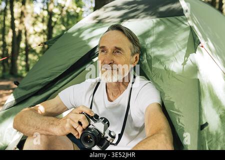Männlicher Tourist mit Filmkamera, der im Wald sitzt. Senior Tourist entspannte sich während seines kleinen Abenteuers in der Nähe des Zeltes Stockfoto