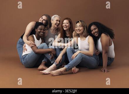 Porträt von sechs lachenden Frauen unterschiedlichen Alters und Körpertypen, die auf braunem Hintergrund im Studio sitzen Stockfoto