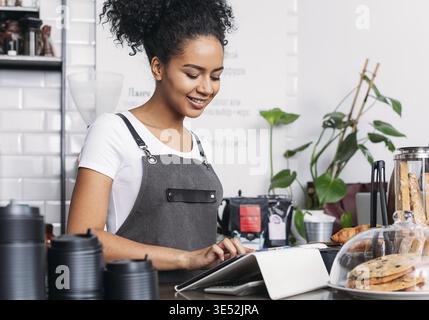 Barista in Schürze auf einem digitalen Tablet. Fröhliche Frau mit lockigem Haar, die als Barista in einem Café mit einem digitalen Tablet arbeitet Stockfoto