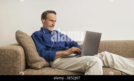 Älterer Mann, der mit einem Laptop auf einer Couch im Wohnzimmer liegt Stockfoto