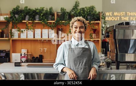 Selbstbewusster Besitzer eines Coffee Shops auf einer Schürze. Junge lächelnde Frau in einer Schürze, die an der Bar steht Stockfoto