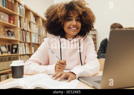 Porträt eines lächelnden jungen Studenten, der in der Bibliothek studiert. Junge Frau am Bücherpult, die in die Kamera schaut Stockfoto
