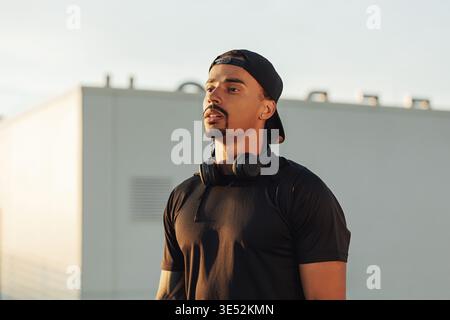 Porträt eines jungen, selbstbewussten männlichen Athleten in einer Mütze. Sportler mit kabellosen Kopfhörern, die bei Sonnenuntergang auf dem Dach stehen Stockfoto