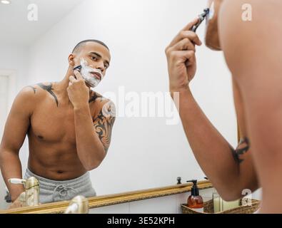 Junger Mann rasiert sich vor einem Spiegel die Wange Stockfoto