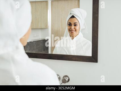 Frau im Bademantel, die ihr Spiegelbild ansieht. Schöne Brünette mit einem weißen Handtuch auf dem Kopf Stockfoto