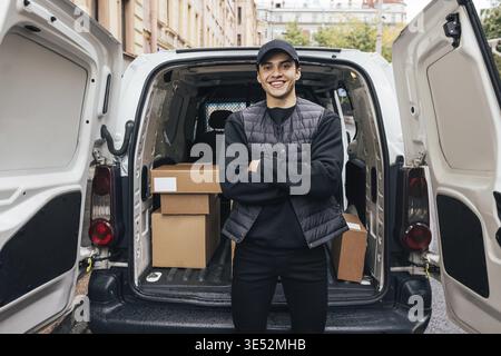 Porträt eines lächelnden Lieferers mit Mütze und Uniform, der mit gekreuzten Armen am Kofferraum des Lieferwagens steht und die Kamera anschaut Stockfoto