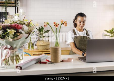 Frau mit Schürze, die in ihrem Blumenladen arbeitet. Junge Geschäftsfrau, die drinnen auf einem Laptop schreibt Stockfoto
