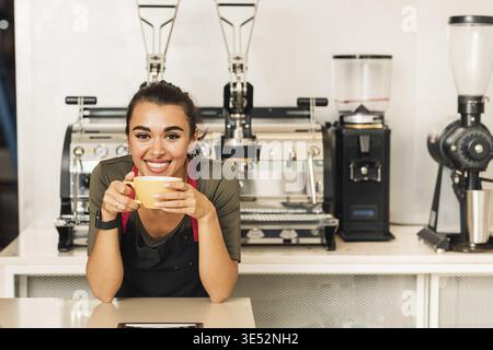 Lächelnde Frau in Schürze, die sich auf einen Tisch stützt, während sie Kaffee trinkt. Porträt einer Kellnerin, die vor der Kaffeemaschine steht Stockfoto