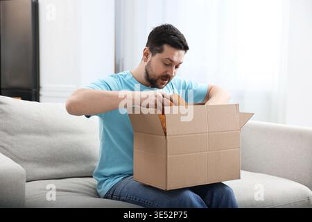 Ein überraschter Mann, der zu Hause das Paket auf dem Sofa öffnet Stockfoto